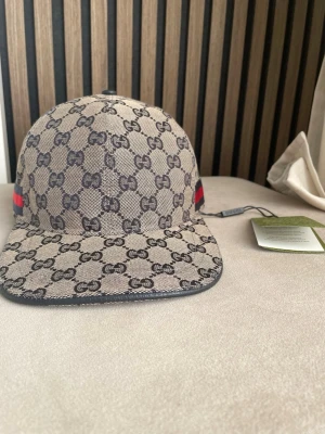 Gucci beige keps med GG-mönster - Säljer en beige keps från Gucci med det klassiska GG-monogrammet över hela kepsen. På sidan finns en ikonisk röd och blå rand. Kepsen har böjd skärm och är tillverkad i canvasmaterial. Perfekt för dig som vill ha en lyxig och trendig accessoar.