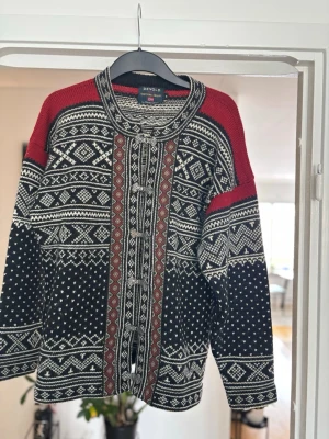 Snygg kofta från Devold - Säljer en traditionell stickad kofta från Devold med snyggt norskt mönster i svart, vitt och vinrött. Koftan har dekorativa knäppningar framtill och rund hals. Perfekt för dig som gillar klassisk och unik stil.
