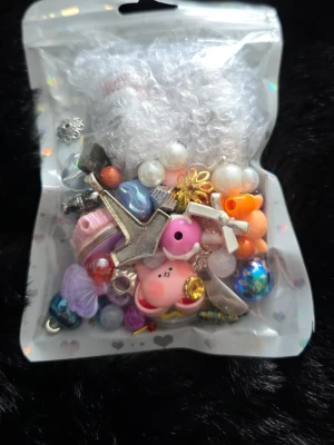 Påse med blandade smyckesdelar - Påse med mixade smyckesdelar och berlocker i olika färger och former, bland annat pärlor, metallberlocker, plastfigurer och dekorativa hängen. Perfekt för DIY och att skapa egna smycken. Innehåller motiv som djur, stjärnor och hjärtan i färgglada material. Även 2 brukar med kristaller chips.