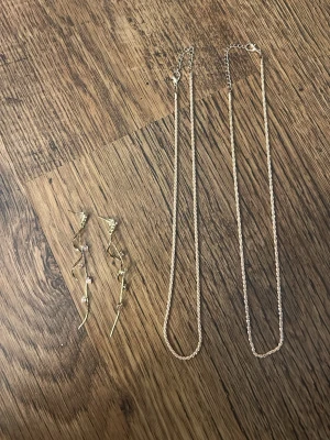 Guldiga hängande örhängen med kristaller halsband - 2 par halsband.Säljer ett par unika guldiga örhängen med vågig form och flera små klara kristaller längs hela längden. Örhängena är långa och ger ett elegant intryck med sina glittrande detaljer. Perfekta för dig som vill sticka ut med accessoarerna. Allt i bilden för 15kr. Aldrig använd