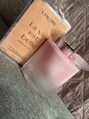 Lancôme La Vie Est Belle Vanille Nude - Lancôme La Vie Est Belle Vanille Nude är en elegant parfym i en mjukt rosa glasflaska med silverfärgad kork och en glittrig rosa rosett runt halsen. Flaskan rymmer 75 ml och har en feminin, modern design som känns lyxig och stilren.