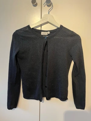 Svart glittrig kofta från KappAhl - Snygg svart kofta från KappAhl med diskret glittrigt material. Rund halsringning, långa ärmar och knäppning framtill med matchande svarta knappar. Perfekt för kalas eller bara det lilla extra🤩! 