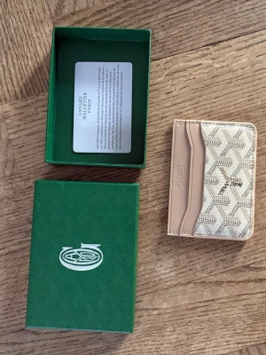 Biege Goyard plånbok - Snyggt och stilrent kortfodral från Goyard i beige och ljusbrunt. Kortfodralet har klassiskt Goyard-mönster, flera kortfack och är tillverkat i läder och canvas. Perfekt för dig som vill ha en exklusiv accessoar i fickan.