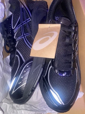 Svarta Asics sneakers med reflexdetaljer - Snygga svarta Asics sneakers med silvriga reflexdetaljer och klassisk mesh- och syntetöverdel. Skorna har en sportig look med tydliga Asics-loggor på sidorna och svart sula. Perfekta för dig som gillar streetwear och vill sticka ut.