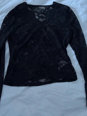 Svart spetsblus med lång ärm - Snygg svart spetsblus med lång ärm och v-ringad hals. Blusen är genombruten i spetsmaterial och har en feminin, figurnära passform. Perfekt att styla med jeans eller kjol för en trendig look.