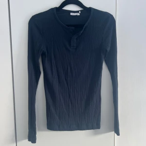ribbad långärmad topp - En mörkblå ribbad långärmad topp med rund halsringning och knappdetalj framtill. Toppen har en slim passform och är tillverkad i mjuk bomullsmix som känns skön mot huden. Perfekt att styla med jeans eller kjol för en enkel och snygg look. Passar s/xs! 