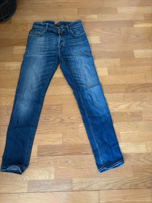 Blå raka jeans från Jacob Cohen  - Säljer ett par klassiska blå jeans från Jacob Cohen med raka ben och snygg slitning. Jeansen har fem fickor, knappgylf och tydliga kontrastsömmar. Perfekta till en avslappnad stil och tillverkade i slitstarkt denimtyg.
