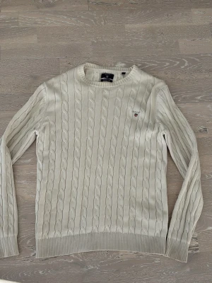 Vit kabelstickad tröja från Gant - Kabelstickad vit tröja från Gant i premium bomull. Klassisk rund halsringning, ribbade muddar och diskret Gant-logga broderad på bröstet. Perfekt för dig som gillar stilrena och tidlösa plagg.
