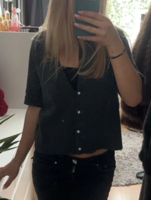 Mörkgrå kortärmad kofta med pärlknappar - Kortärmad mörkgrå kofta med v-ringning och dekorativa pärlknappar framtill. Modellen är något croppad och har en avslappnad passform. Perfekt att styla över ett linne eller t-shirt för en trendig lager-på-lager-look.