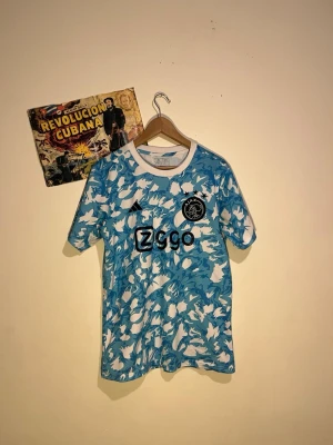 Ajax blåvit matchtröja Adidas - Ajax matchtröja i blått och vitt camomönster med svart Ajax-logga och Ziggo sponsortryck på bröstet. Tröjan har rund hals med vit krage och är tillverkad i lätt och ventilerande material. Perfekt för fotbollsträning eller att bära som supporter.