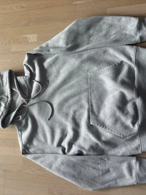 Grå hoodie från Tommy Hilfiger - Snygg grå hoodie från Tommy Hilfiger med liten logga på bröstet. Klassisk känguruficka framtill, dragsko i huvan och ribbade muddar vid ärmar och nederkant. Tillverkad i mjuk bomullsblandning som känns skön mot huden. Sitter lite med som en M än L