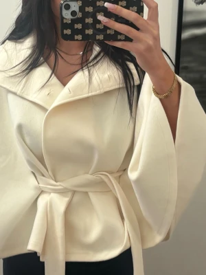 Krämvit kappa med knytskärp - Stilren krämvit kappa med bred krage och knytskärp i midjan. Kappan har vida ärmar och en mjuk, slät yta som ger ett elegant intryck. Perfekt för dig som gillar minimalistisk och modern stil.
