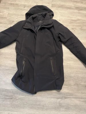 Marinblå UBR jacka Parka regulator - Nypris: 9000kr  I  Mitt pris: 5999kr                Stilren svart parkasjacka från UBR med huva och två snedställda dragkedjefickor framtill. Jackan har en rak passform och diskret logga på bröstet. Materialet är vattenavvisande och passar perfekt för kyliga dagar. Passar dig som är mellan 170-185cm lång