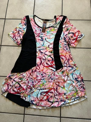 Färgglad kortklänning från Nareed - Unik kortklänning från Nareed med färgstarkt abstrakt mönster i rosa, rött, blått och grönt. Svarta paneler längs sidorna ger en cool kontrast. Klänningen har rund halsringning och korta ärmar. Perfekt för dig som gillar statement-plagg!