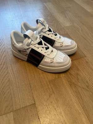 Vita Valentino sneakers med svart detalj - Snygga vita sneakers från Valentino med svart panel på sidan och röda VLTN-detaljer framtill. Skorna har klassisk låg profil, perforerad tå och vita skosnören. Tillverkade i läder med stilren design och tydlig logga på plösen.