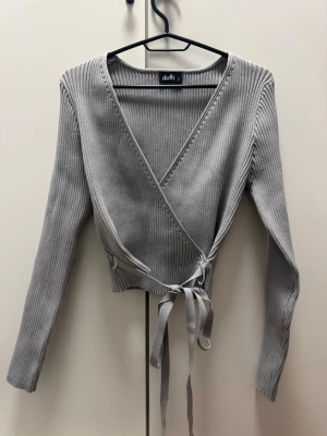 Grå omlottopp från Dotti - Snygg grå ribbad omlottopp från Dotti med långa ärmar och knytband i midjan. Tröjan har en djup v-ringning och är tillverkad i ett stretchigt material som sitter tajt och bekvämt. Nyskick