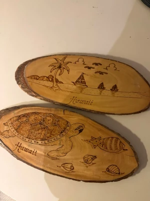 Dekorativa träplattor Hawaii-motiv - Två ovala träplattor med ingraverade Hawaii-motiv. Ena plattan har en havssköldpadda och snäckor, den andra visar en strand med palmer, segelbåt och fåglar. Båda har texten 'Hawaii' ingraverad. Naturfärgat trä med barkkant.