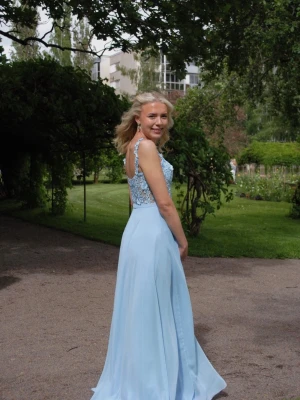 Ljusblå långklänning med spetsdetaljer - Elegant ljusblå långklänning med spetsdetaljer upptill och smala axelband. Klänningen har v-ringad urringning, omlott och slits framtill för extra rörelsefrihet. Kjoldelen är i ett lätt, svepande material som ger en romantisk känsla.
