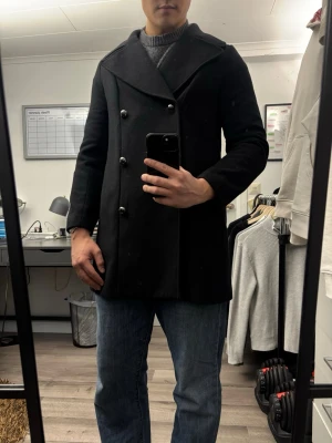 Svart dubbelknäppt rock - Stilren svart rock med dubbelknäppning och bred krage. Klassisk modell med sex knappar framtill och slät yta. Passar perfekt till jeans eller mer uppklätt. Troligen i ullblandning med slätt foder på insidan.