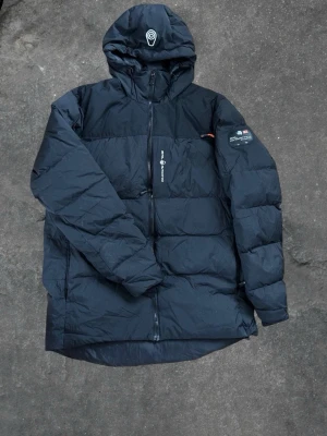 Svart pufferjacka från Sail Racing - Säljer en svart pufferjacka från Sail Racing med huva och dragkedja framtill. Jackan har en patch på ärmen och logga på huvan. Fylld med vaddering för extra värme och har en sportig, oversized look. Perfekt för kalla vinterdagar.