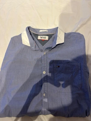 Blårandig skjorta från Tommy Hilfiger - Snygg blårandig skjorta från Tommy Hilfiger med vit kontrastkrage och diskret logga på bröstfickan. Skjortan har långa ärmar, knappar framtill och är gjord i mjuk bomull. Perfekt för en clean och stilren look.