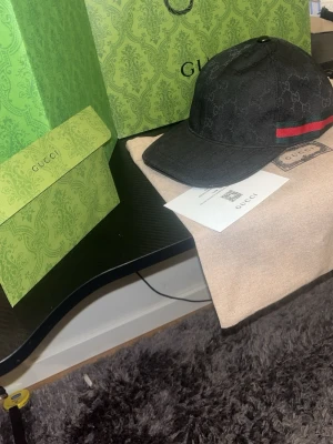 Svart Gucci keps med GG-mönster - Säljer en svart keps från Gucci med det klassiska GG-monogrammönstret över hela kepsen. På sidan finns den ikoniska gröna och röda randen. Kepsen är tillverkad i canvasmaterial och har böjd skärm. Perfekt för dig som vill ha en exklusiv och stilren accessoar.