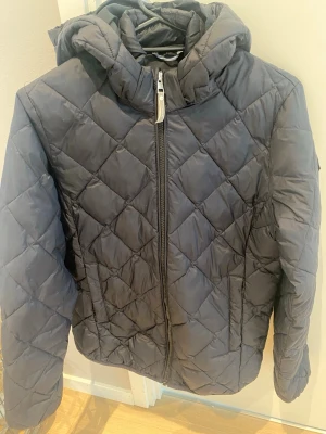 Mörk blå pufferjacka från Gant - Säljer en mörk blå pufferjacka från Gant med quiltat mönster och huva. Jackan har dragkedja framtill, två sidofickor en innerficka och en snygg Gant-logga på ärmen. Perfekt till vår och sommaren och riktigt skön att bära.