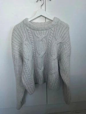 Vit/creme stickad tröja - Säljer en supermysig  stickad tröja med grova flätstickade detaljer och ribbade muddar. Tröjan har en relaxed fit och långa ärmar, perfekt för kyliga dagar. Passar dig som gillar en oversized och trendig look.