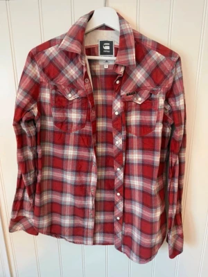 Rutig röd skjorta från G-Star Raw - Säljer en röd och vit rutig skjorta från G-Star Raw i storlek M. Skjortan har långa ärmar, klassisk krage, tryckknappar och två bröstfickor. Tillverkad i mjuk bomull, perfekt för en avslappnad och cool stil.
