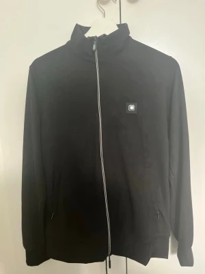 Svart dress  från Calvin Klein - Svart dress från Calvin Klein med hög krage och dragkedja framtill. Tröjan har en liten CK-logga på bröstet och ribbad struktur i materialet. Enkel och stilren design som funkar till det mesta. (Tröja& byxor)