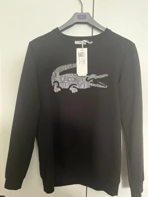 Svart tröja från Lacoste - Svart tröja  från Lacoste med stort Lacoste märke på bröstet Tillverkad i mjukt material som passar perfekt till en avslappnad stil.