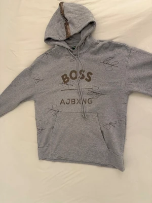 Grå hoodie från BOSS AJBXNG - Snygg grå hoodie från BOSS AJBXNG med brunt tryck framtill och signaturmönster över hela plagget. Hoodien har en huva med brunt band, dragsko och en stor magficka. Tillverkad i mjukt sweatshirtmaterial, perfekt för en avslappnad streetwear-look.