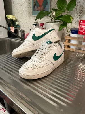 Vita Nike sneakers med gröna detaljer - Säljer ett par klassiska vita Nike sneakers med gröna swoosh-loggor på sidorna och matchande grönt Nike-märke på plösen. Skorna har perforerad tå för ventilation och är tillverkade i läder med lågt skaft och vita snören. Perfekta för en clean och sportig look. Säljer dom för att dom är för liten för mig. Jag köpte den från zalando för 980kr. Den är bra skick och har ingen skador, bara att den är smutsig.