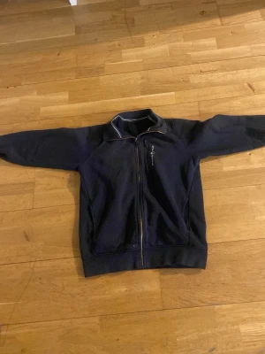 Svart hoodie från Sail Racing - Svart hoodie från Sail Racing med dragkedja framtill och en extra bröstficka med dragkedja. Stilig ståkrage med vit detaljsöm och broderad logga på bröstet. Tillverkad i mjukt bomullsmaterial, perfekt för en avslappnad och sportig stil. Pris kan diskuteras.