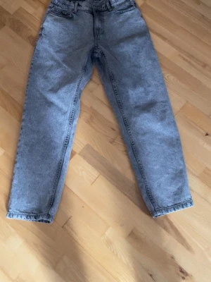 Grå mom jeans med hög midja - Säljer ett par grå mom jeans med hög midja och raka ben. Jeansen har klassisk femficksdesign och är tillverkade i ett mjukt denimtyg. Perfekta för en avslappnad och trendig look.