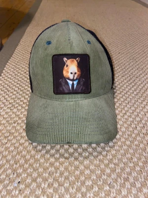 Olivgrön truckerkeps med djurpatch - Säljer en olivgrön truckerkeps med svart mesh baktill och justerbart spänne. Framtill sitter en patch med en capybara i kostym. Kepsen är i manchestertyg framtill och har en unik, lekfull design.