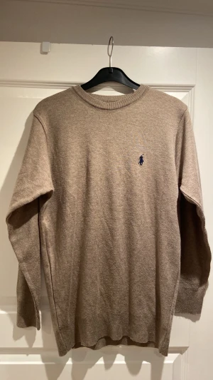 Beige tröja från Ralph Lauren - Klassisk beige tröja från Ralph Lauren med rund halsringning och den ikoniska mörkblå broderade loggan på bröstet. Tröjan är stickad i mjukt material och har ribbade muddar vid ärmslut och nederkant. Perfekt för en stilren och avslappnad look.pris är diskuterbart 