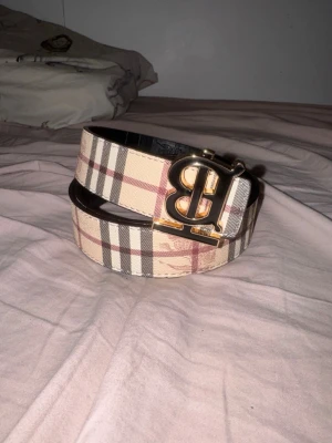 Burberry bälte med klassiskt rutmönster - Säljer ett Burberry bälte med ikoniskt rutmönster i beige, svart och rött samt en stor guld B-spänne. Bältet är tillverkat i skinn och har en exklusiv känsla. Perfekt accessoar för att lyfta din outfit.