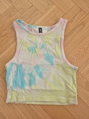 Pastellfärgad ribbad linnetopp H&M - Säljer en ribbad linnetopp från H&M Divided i storlek XS. Toppen har ett pastellfärgat batikmönster i blått, gult och rosa samt är ärmlös och croppad. Perfekt för varma dagar och enkel att styla med jeans eller shorts.