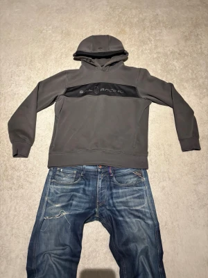 Grå hoodie från Sail Racing - Snygg grå hoodie från Sail Racing med svart panel och logga över bröstet. Klassisk huva med dragsko och ribbade muddar vid ärmslut och nederkant. Tillverkad i mjukt material som känns skönt mot huden. Perfekt för en avslappnad och sportig stil. Mycket volym i luvan så att den inte säckar ihop