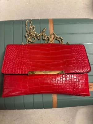 Röd kuvertväska från Dune London - Snygg röd kuvertväska från Dune London i croco-mönstrat konstläder. Väskan har guldfärgad kedja och metallist med logga framtill. Perfekt för dig som vill sticka ut med en färgstark accessoar.
