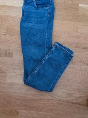 Blå skinny jeans i klassisk modell 152 - Säljer ett par blå skinny jeans med hög midja och smal passform. Jeansen har klassiska fem fickor och är gjorda i stretchigt denimtyg för extra komfort. Perfekta till en avslappnad look och lätta att matcha med olika stilar.