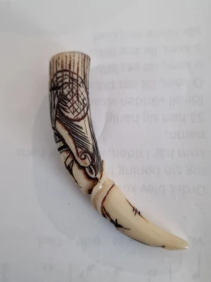 Halsbandshänge i form av horn - Unikt halsbandshänge format som ett horn med inristade mönster och detaljer. Färgen är benvit med mörkbruna gravyrer som ger en cool, rustik känsla. Perfekt för dig som gillar naturinspirerade accessoarer och vill sticka ut.