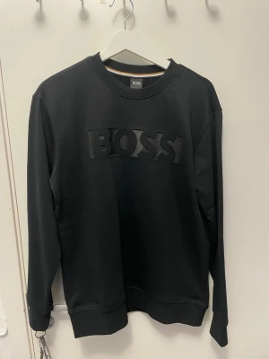 Svart sweatshirt från BOSS - Säljer en svart sweatshirt från BOSS med stor, upphöjd logga framtill. Tröjan har rund halsringning, långa ärmar och ribbade muddar vid ärmslut och nederkant. Tillverkad i mjukt material som känns skönt mot huden. Perfekt för en clean och stilren look.
