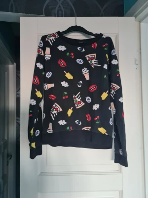 Svart stickad tröja med färgglada tryck - Cool svart stickad tröja med färgglada tryck av pizza, glass, körsbär, läppar och ögon. Tröjan har rund halsringning och långa ärmar. Perfekt för dig som gillar lekfulla och unika plagg med mycket personlighet.