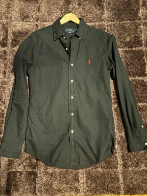 Svart skjorta från Polo Ralph Lauren - Klassisk svart skjorta från Polo Ralph Lauren med röd broderad logga på bröstet. Skjortan har lång ärm, knappar framtill och en tidlös krage. Tillverkad i bomull för en bekväm känsla och snygg passform.
