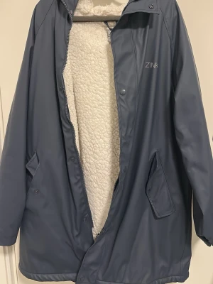 Blå regnjacka med foder från Zink - Snygg blå regnjacka från Zink i storlek M. Jackan har ett mjukt vitt teddyfoder på insidan, dragkedja och tryckknappar framtill samt två snedställda fickor. Perfekt för regniga dagar när du vill hålla dig både torr och varm.