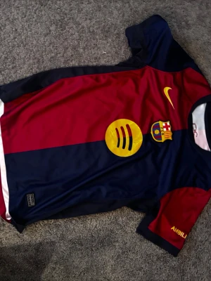 FC Barcelona matchtröja Nike röd/blå - Säljer en FC Barcelona matchtröja från Nike i rött och marinblått med gula detaljer. Tröjan har klubbmärke, Nike-logga och sponsortryck på bröstet samt namn och nummer (Lamine Yamal 19) på ryggen. Tillverkad i lätt och ventilerande material, perfekt för fotboll.