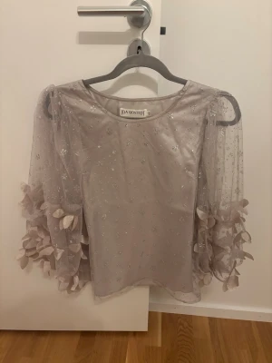 Glittrig topp från Ida Sjöstedt - Superfin ljusgrå/silvrig topp från Ida Sjöstedt med transparenta, glittriga ärmar och blommiga detaljer. Toppen har en rak passform och är täckt av små silverglittrande prickar. 💖Använd en gång
