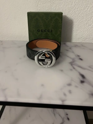 Svart GG Supreme bälte från Gucci - Snyggt svart bälte från Gucci med klassiskt GG Supreme-mönster och stor silverfärgad GG-logga som spänne. Bältet har en mörkgrå ton och insidan är ljusbrun. Perfekt accessoar för att lyfta din outfit.